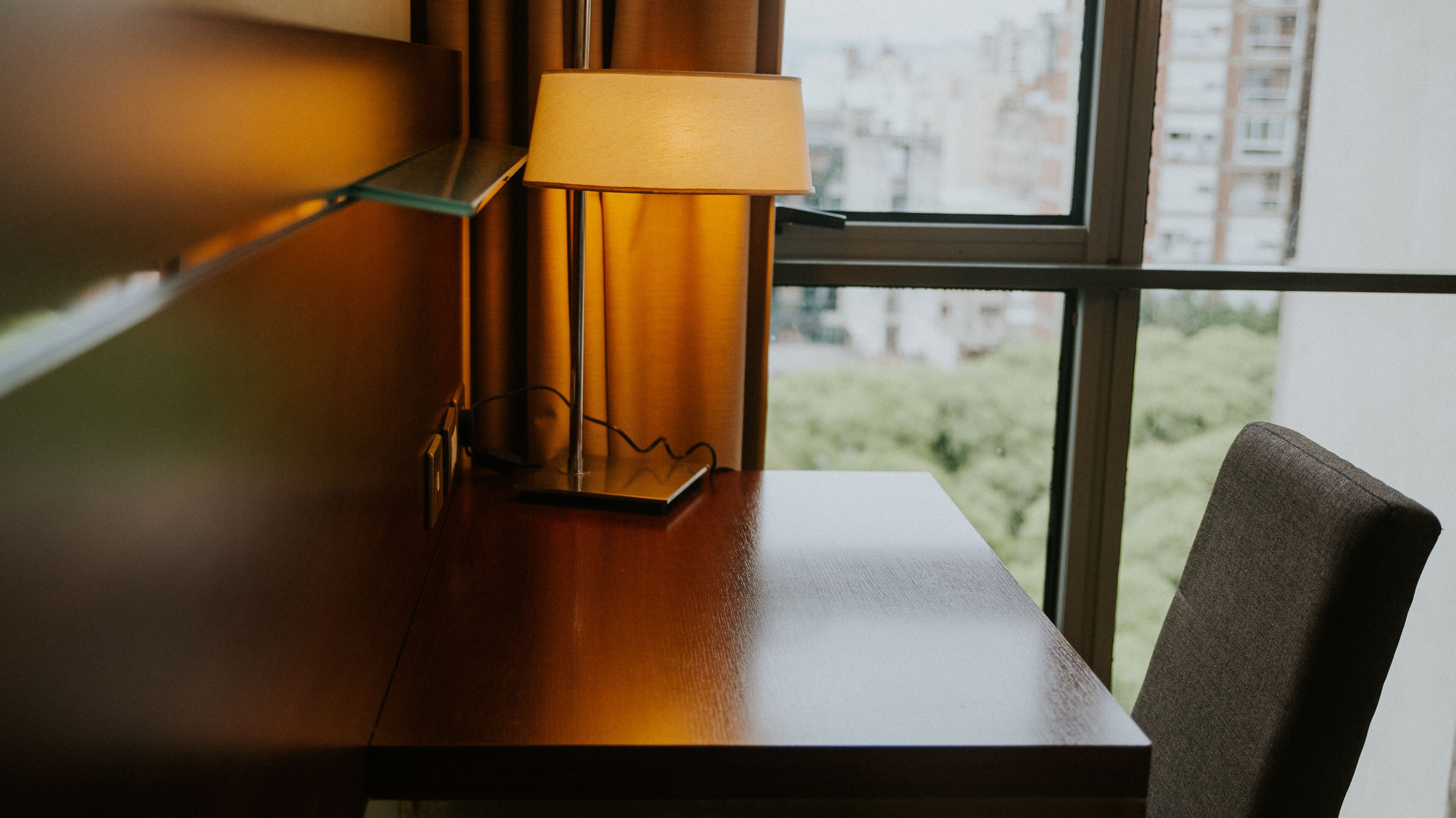 A wooden table with a lamp and a chair by a window with a view of buildings.
