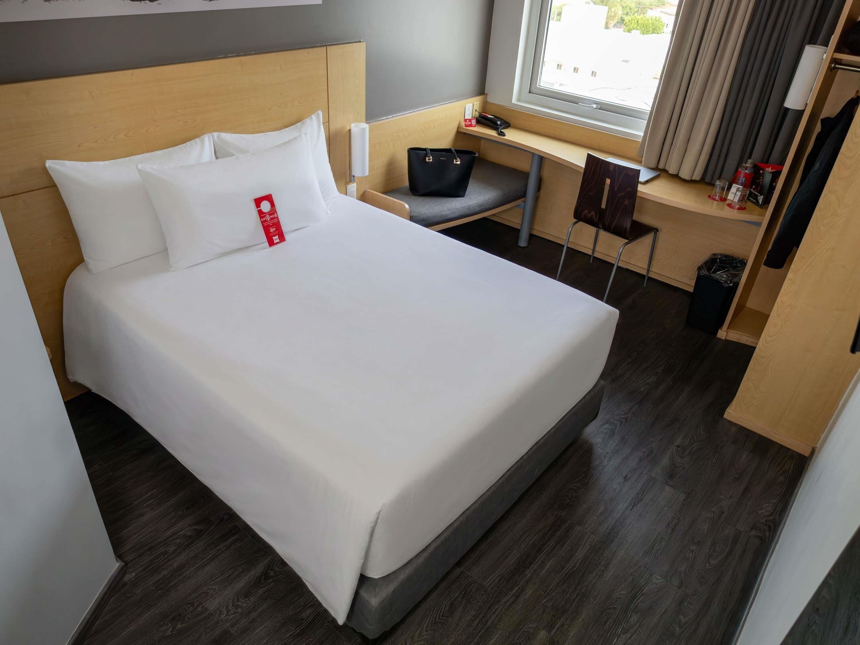 Una cama bien hecha con ropa de cama blanca, cabecera de madera, un escritorio pequeño con silla, un cubo de basura y una ventana con cortinas.