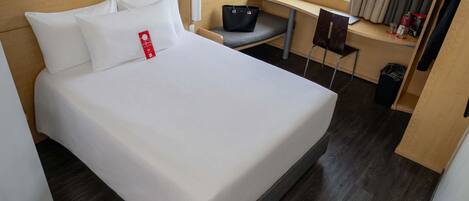 Una cama bien hecha con ropa de cama blanca, cabecera de madera, un escritorio pequeño con silla, un cubo de basura y una ventana con cortinas.