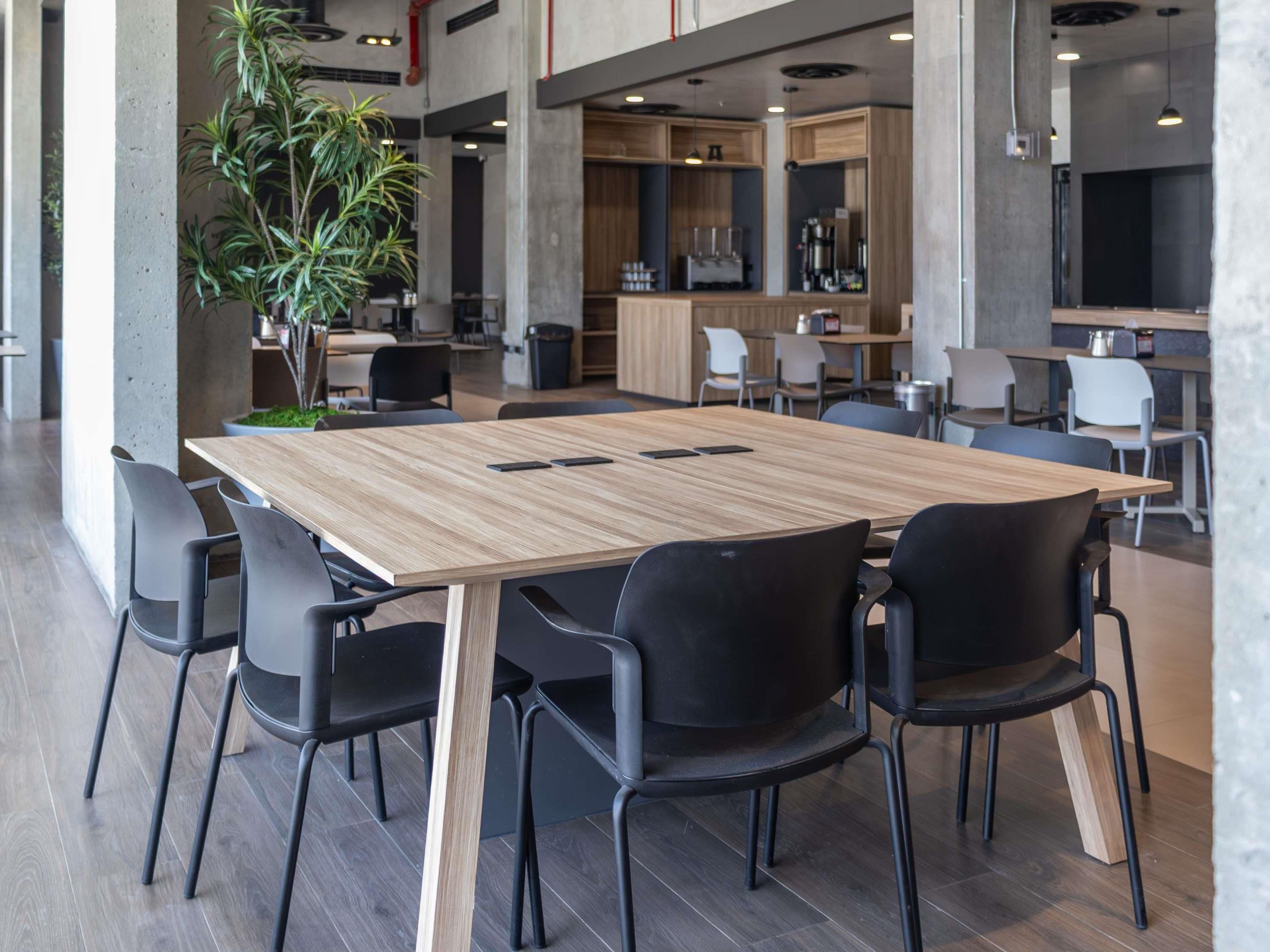 A modern conference room with a wooden table and black chairs, surrounded by a kitchen area with barista equipment.