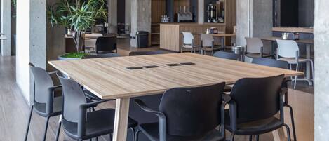 A modern conference room with a wooden table and black chairs, surrounded by a kitchen area with barista equipment.