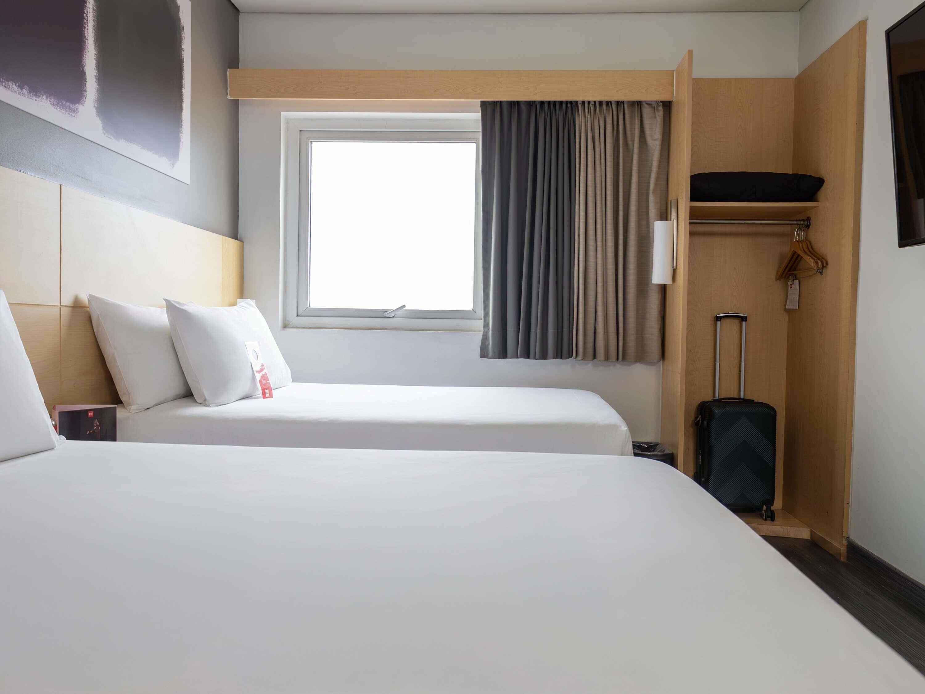 A hotel room with two beds, a window with curtains, a wooden shelf, and a suitcase.