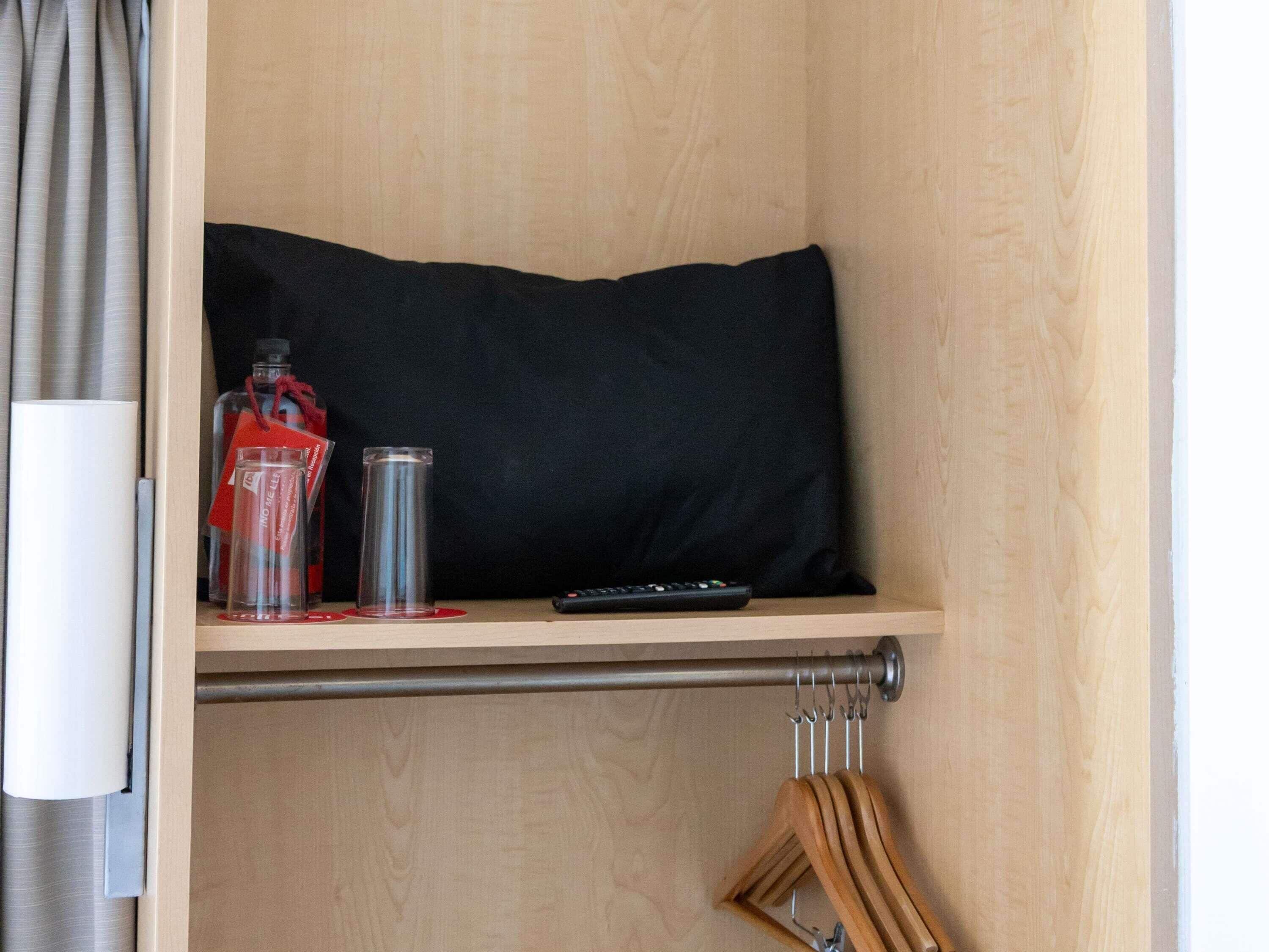 A wooden shelf with a black pillow, a remote control, and a glass bottle with a red tag.