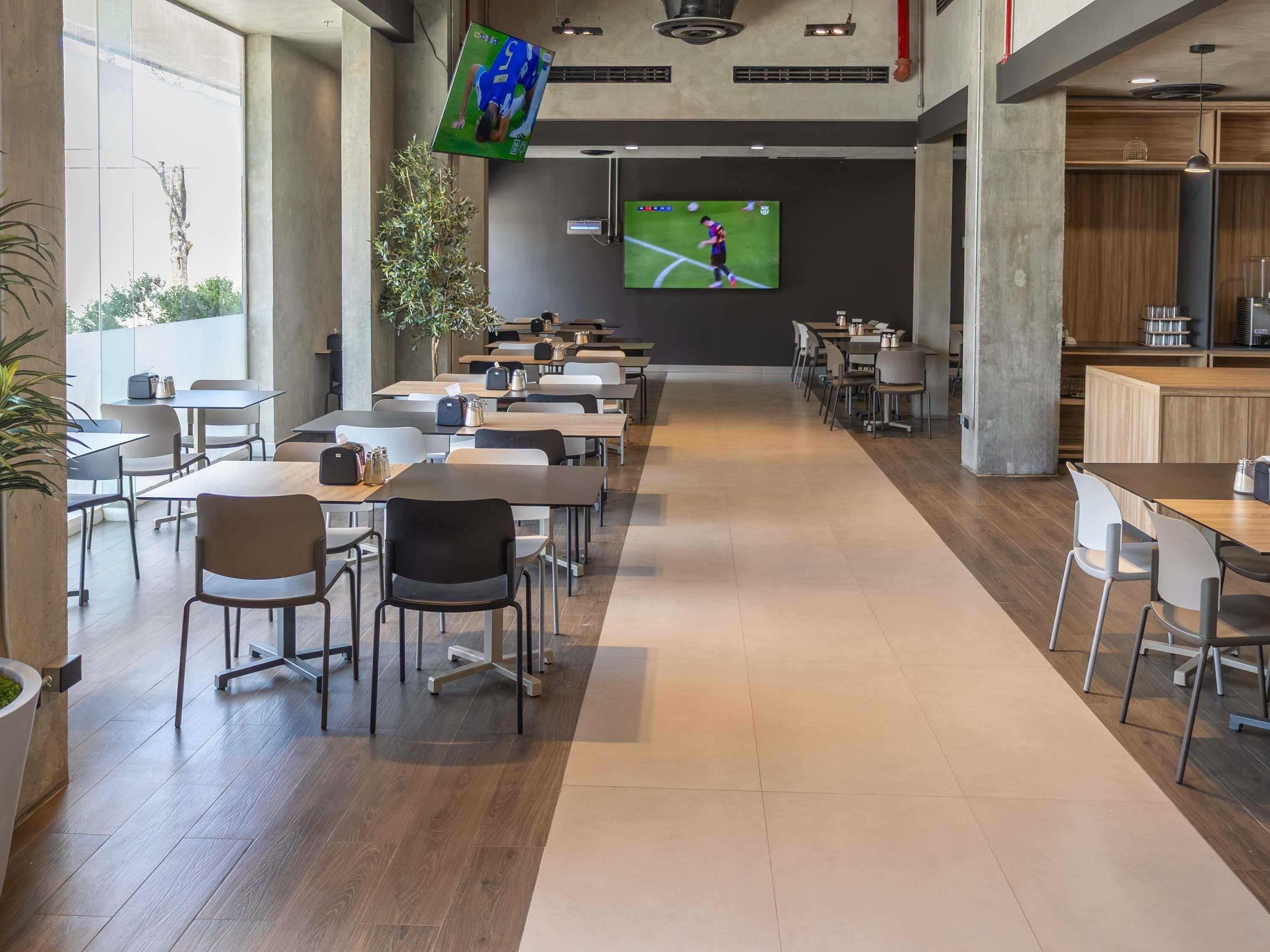 A modern dining area with multiple tables and chairs, a large screen displaying a sports event, and a plant in the center.