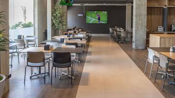 A modern dining area with multiple tables and chairs, a large screen displaying a sports event, and a plant in the center.