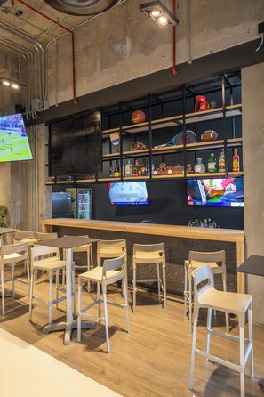 A bar area with wooden chairs and tables, shelves with bottles and sports memorabilia, and a television screen displaying a sports event. - ibis Hermosillo (Hermosillo)