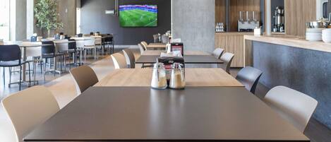 A modern dining area with a long table, multiple chairs, a television screen, and a kitchen counter.