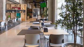 Una cafetería moderna con mesas y sillas largas, un gran árbol en maceta y un techo visible con conductos.