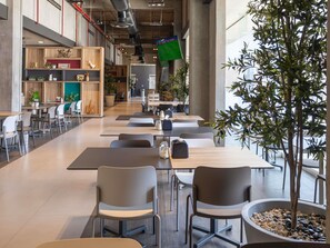A modern cafeteria with long tables and chairs, a large potted tree, and a visible ceiling with ductwork.