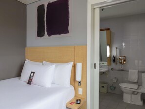 A hotel room with a bed, a bedside table, a nightstand, a wall-mounted lamp, and a bathroom visible through an open door. - ibis Hermosillo (Hermosillo)
