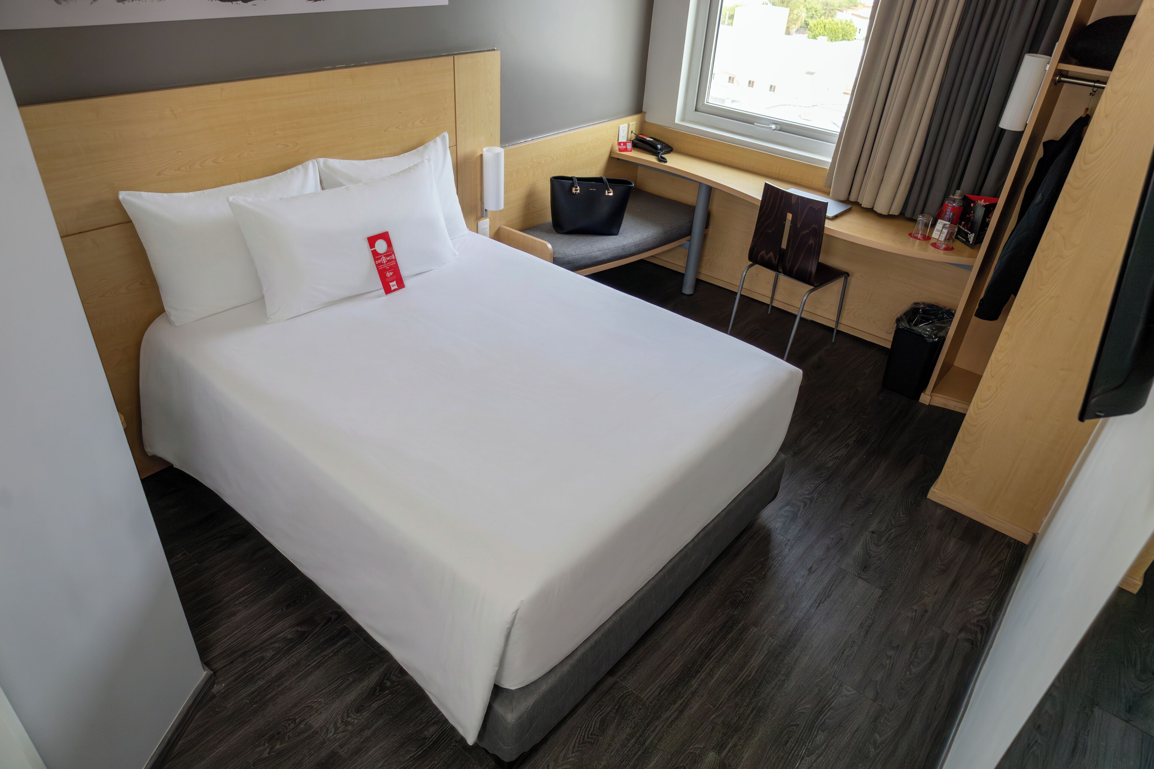 A hotel room with a bed, a desk, a chair, a small table, a trash bin, and a window with curtains.