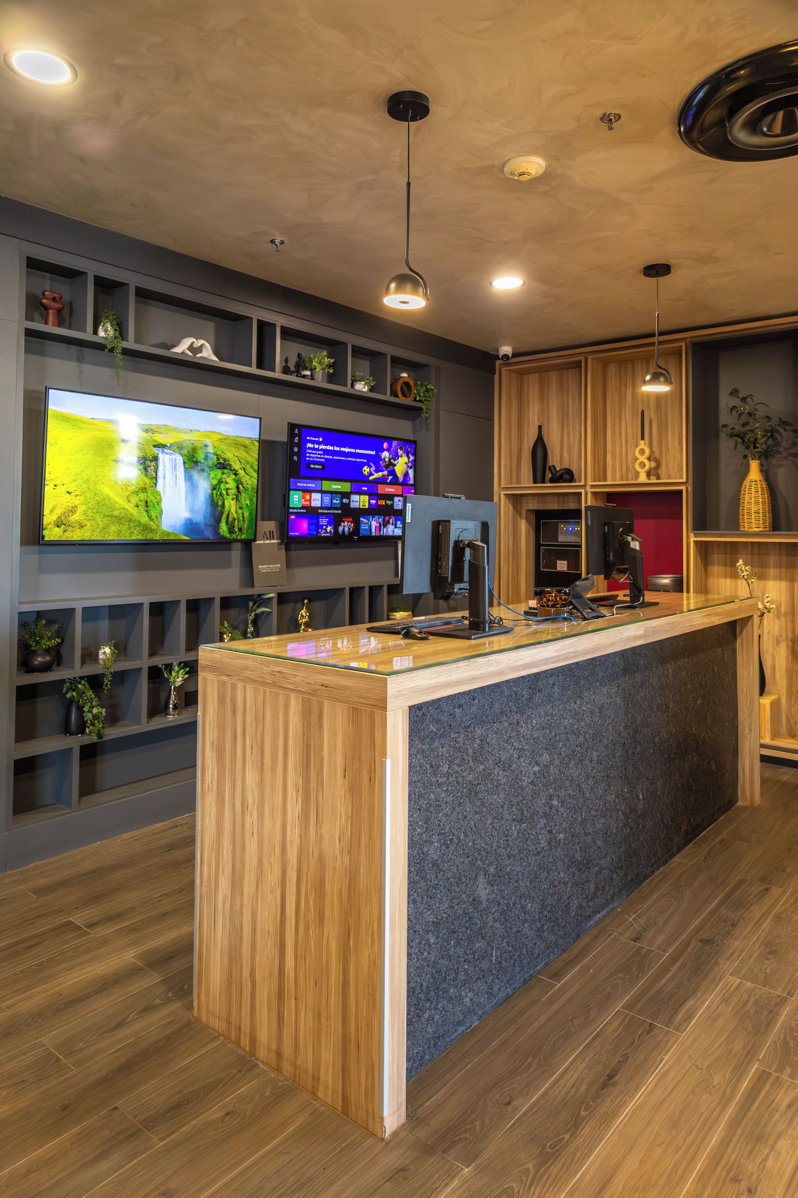 A reception area with a wooden reception desk, a flat-screen TV displaying a nature scene, a digital screen with various icons, and shelves with decorative items.