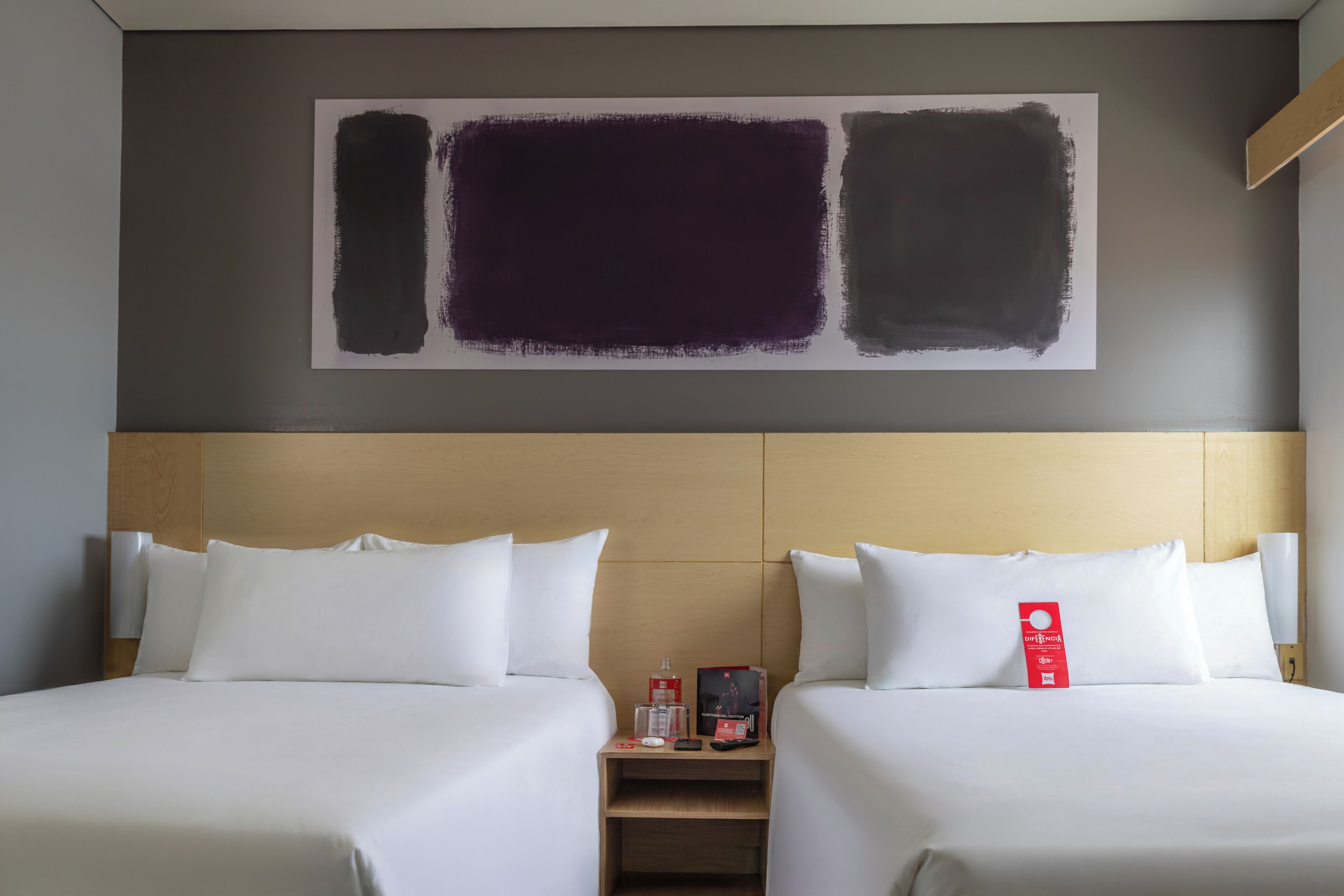 Habitación de hotel con dos camas, cabecera de madera, mesita de noche con un letrero rojo y un cuadro abstracto grande en la pared.