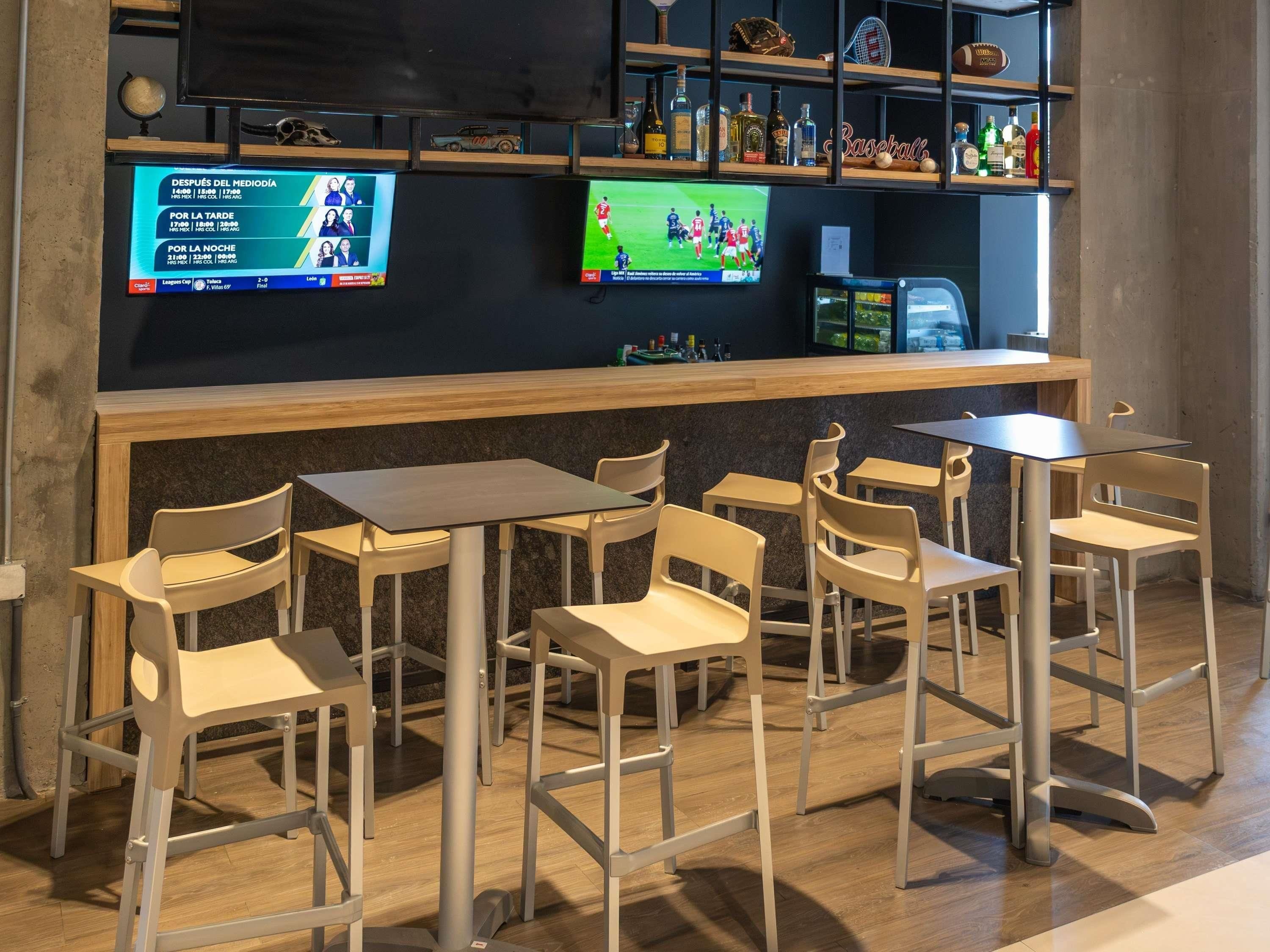 Zona de bar con diversas opciones de asientos, incluyendo mesas y sillas, y una pantalla mostrando contenido deportivo.