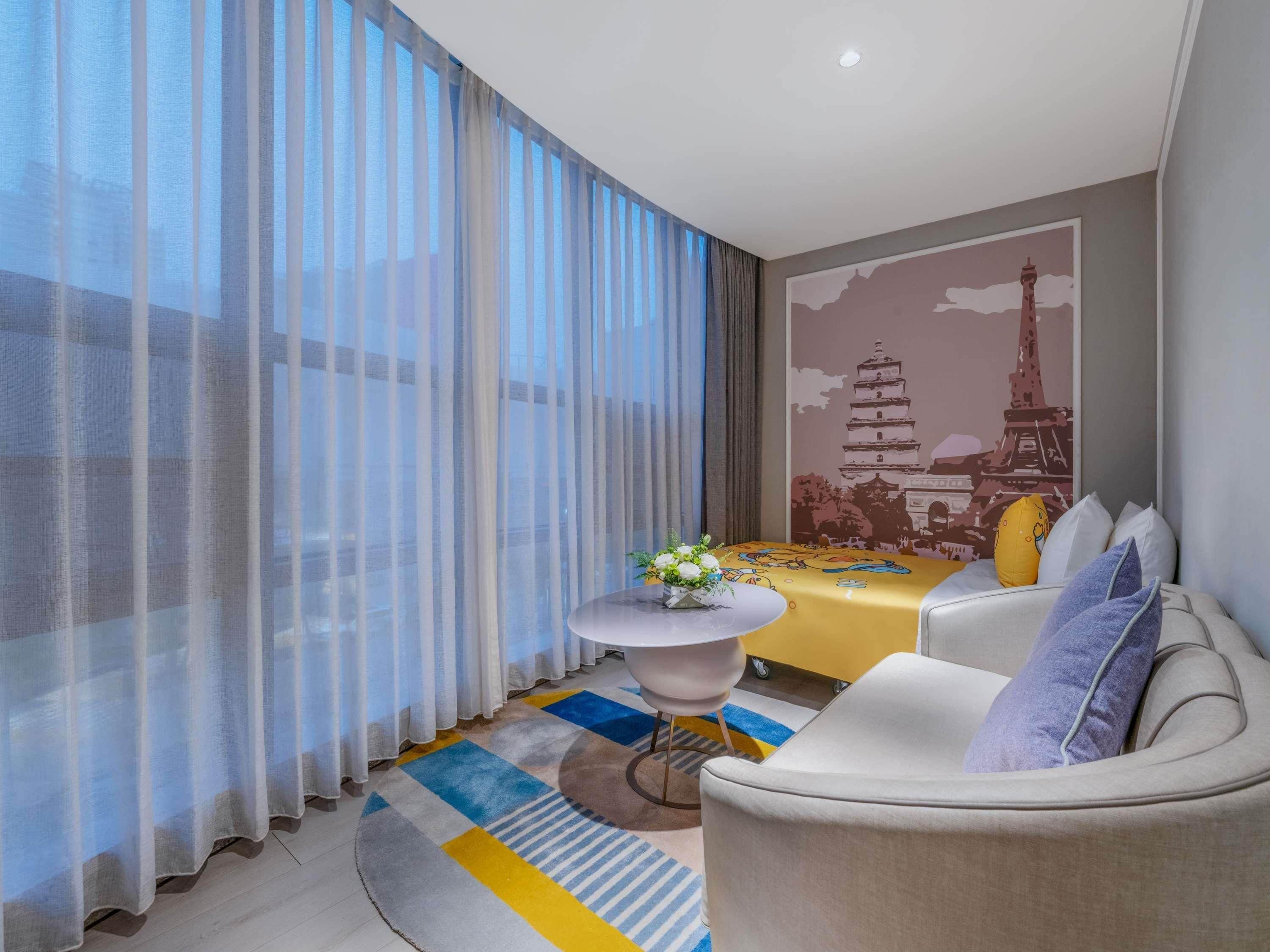 A modern hotel room with a dining table, a sofa, a small round table with a vase, sheer curtains, and a wall mural.