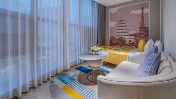 A modern hotel room with a dining table, a sofa, a small round table with a vase, sheer curtains, and a wall mural.