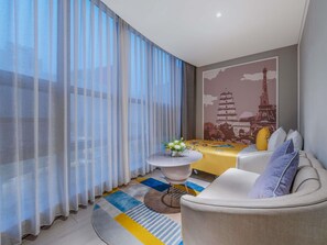 Una habitación de hotel moderna con una mesa de comedor, un sofá, una pequeña mesa redonda con un jarrón, cortinas de gasa y un mural en la pared.