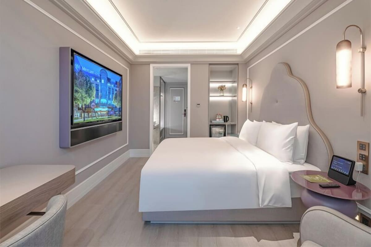 A modern hotel room with a large bed, a flat-screen TV mounted on the wall, a bedside table with a lamp, and a small desk with a chair.