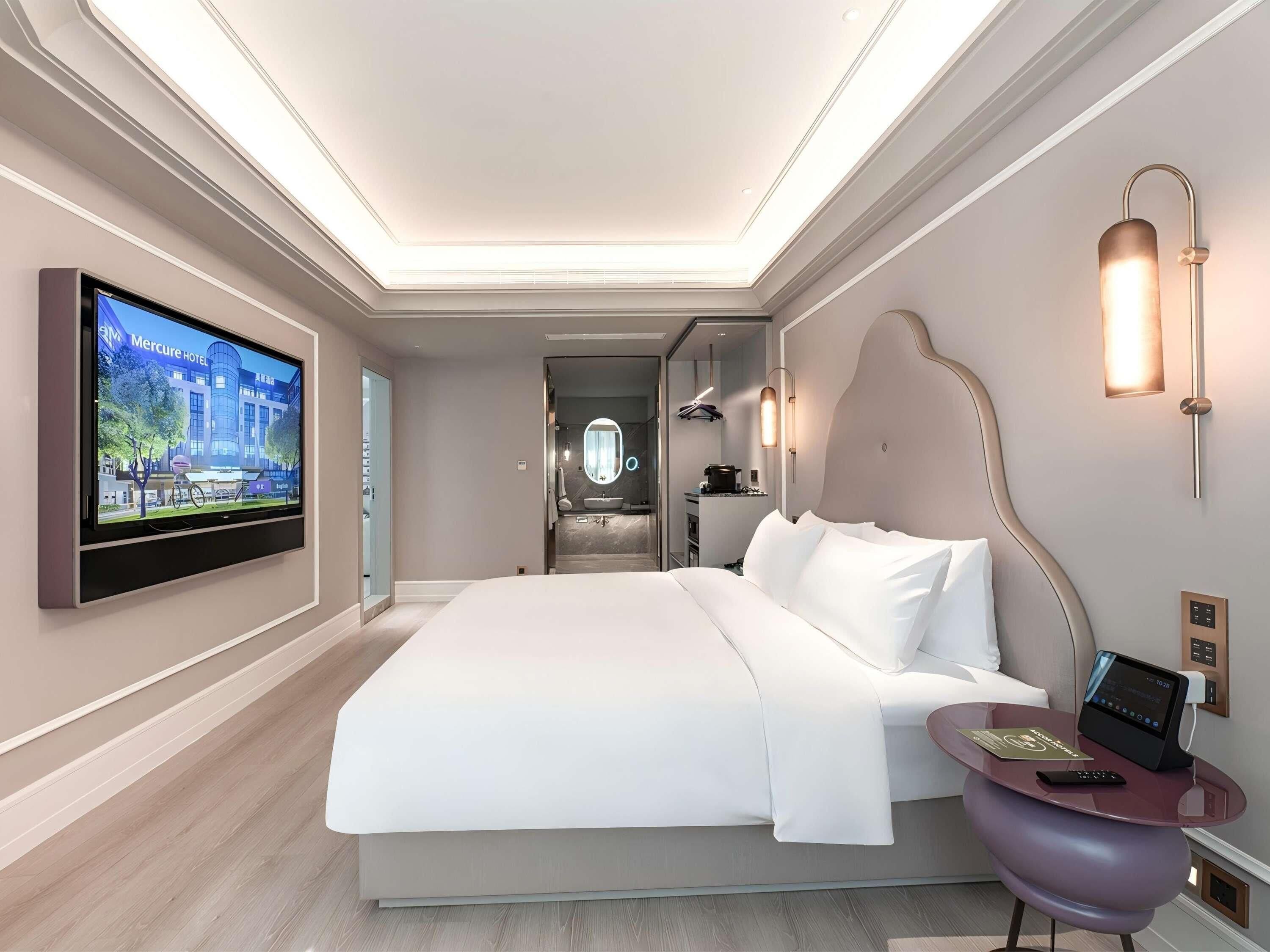 A modern hotel room with a large bed, a television mounted on the wall, a small table with a remote control, and a bathroom visible through an open door.