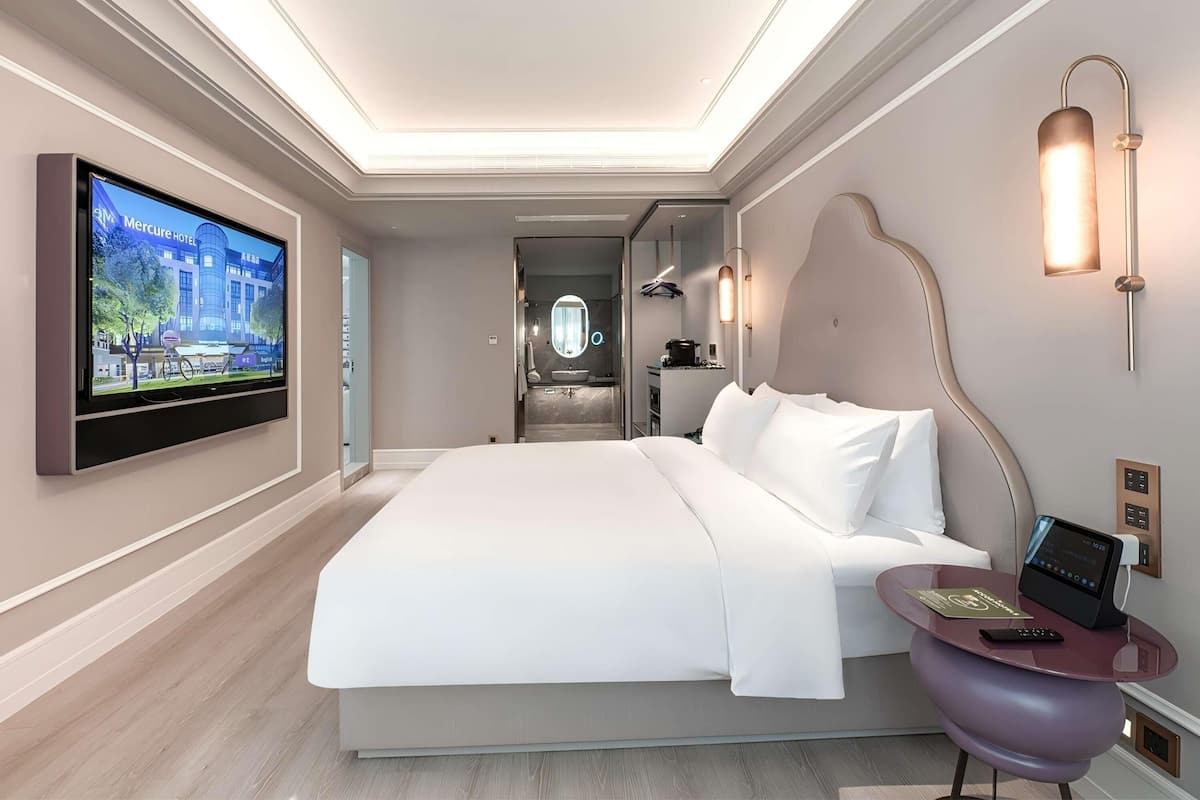 A modern hotel room with a large bed, a television mounted on the wall, a small table with a remote control, and a bathroom visible through an open door.