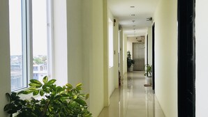 A long, narrow hallway with white walls and a shiny tiled floor. There are large windows on one side, allowing natural light to enter. The hallway leads to a door at the end. - Canary Hotel Hue (Hue)