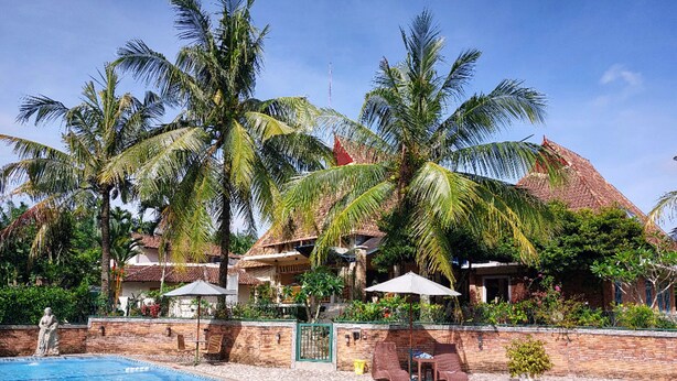 A swimming pool with lounge chairs and umbrellas, surrounded by palm trees and buildings with red-tiled roofs. - Hotel Deli River (Medan)