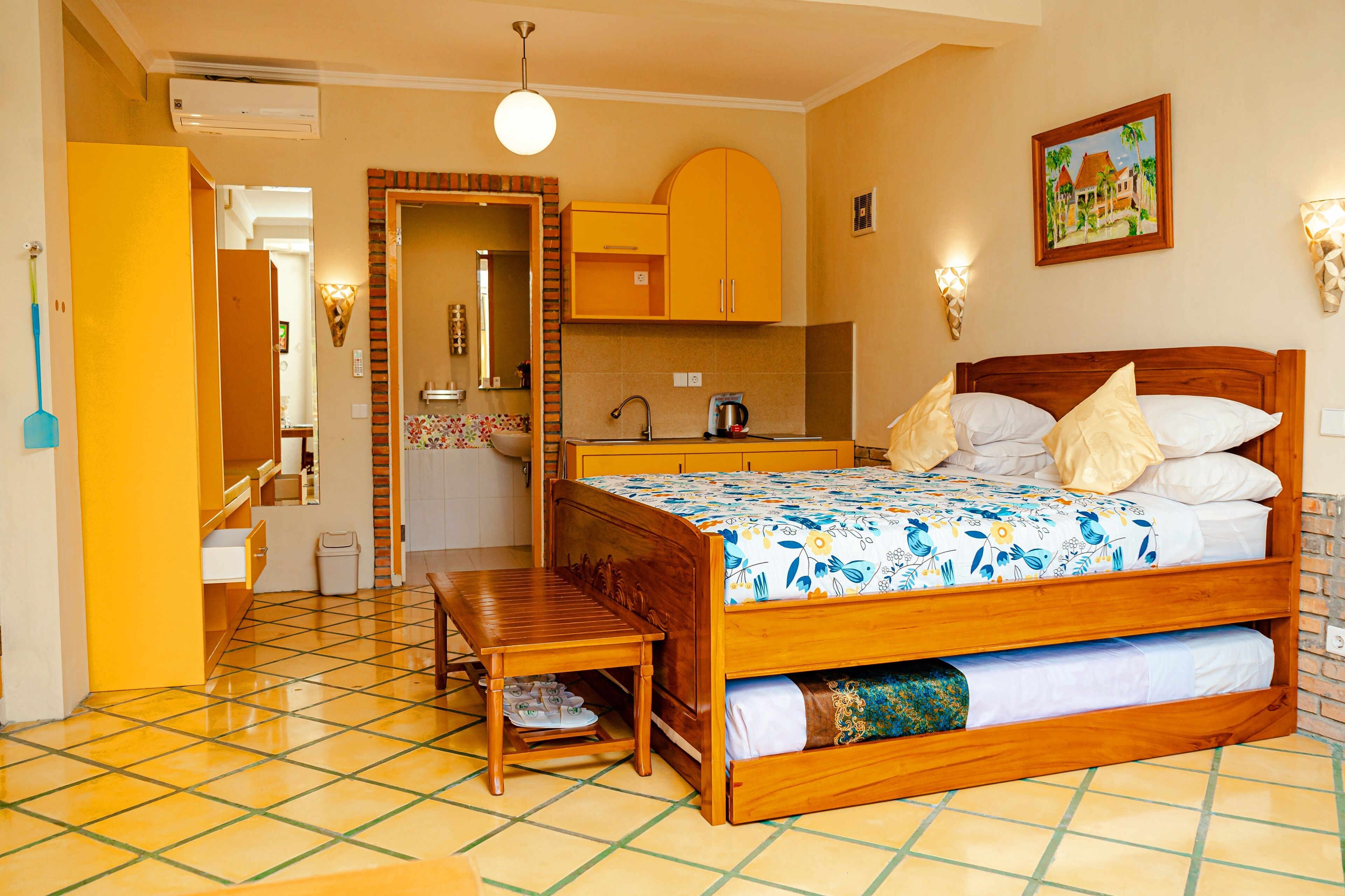 A hotel room with a wooden bed, a small table, a bench, a kitchen area, and a bathroom.