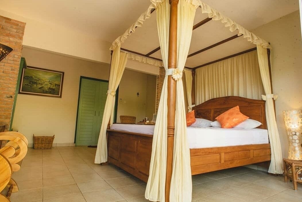 A four-poster canopy bed with a wooden headboard and pillows, a green door, a wicker chair, and a framed picture on the wall.