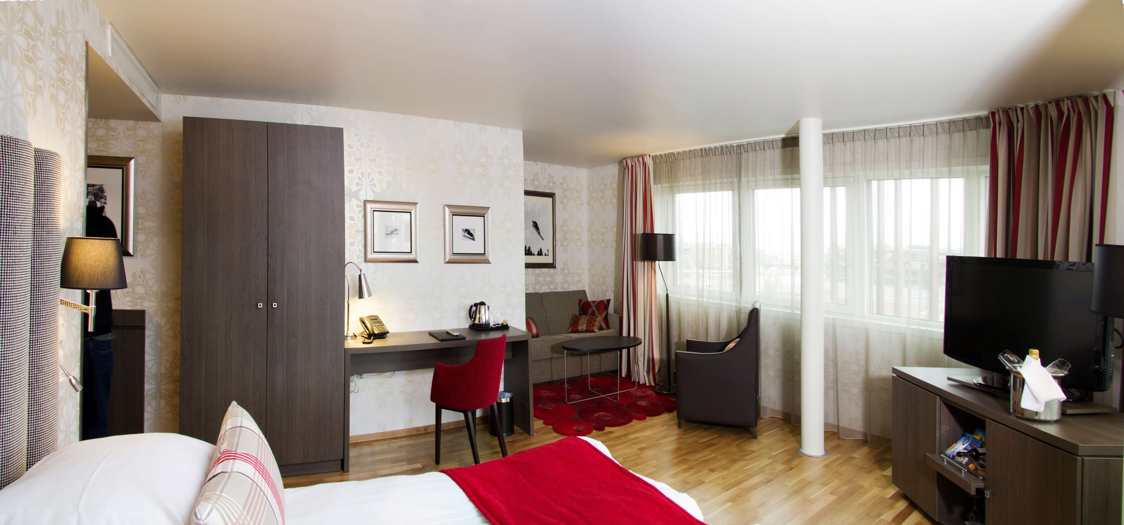 A hotel room with a bed, a desk with a chair, a TV, and a window with curtains.