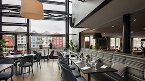 A modern restaurant with a long dining area, black upholstered seating, and large windows offering a view of buildings.