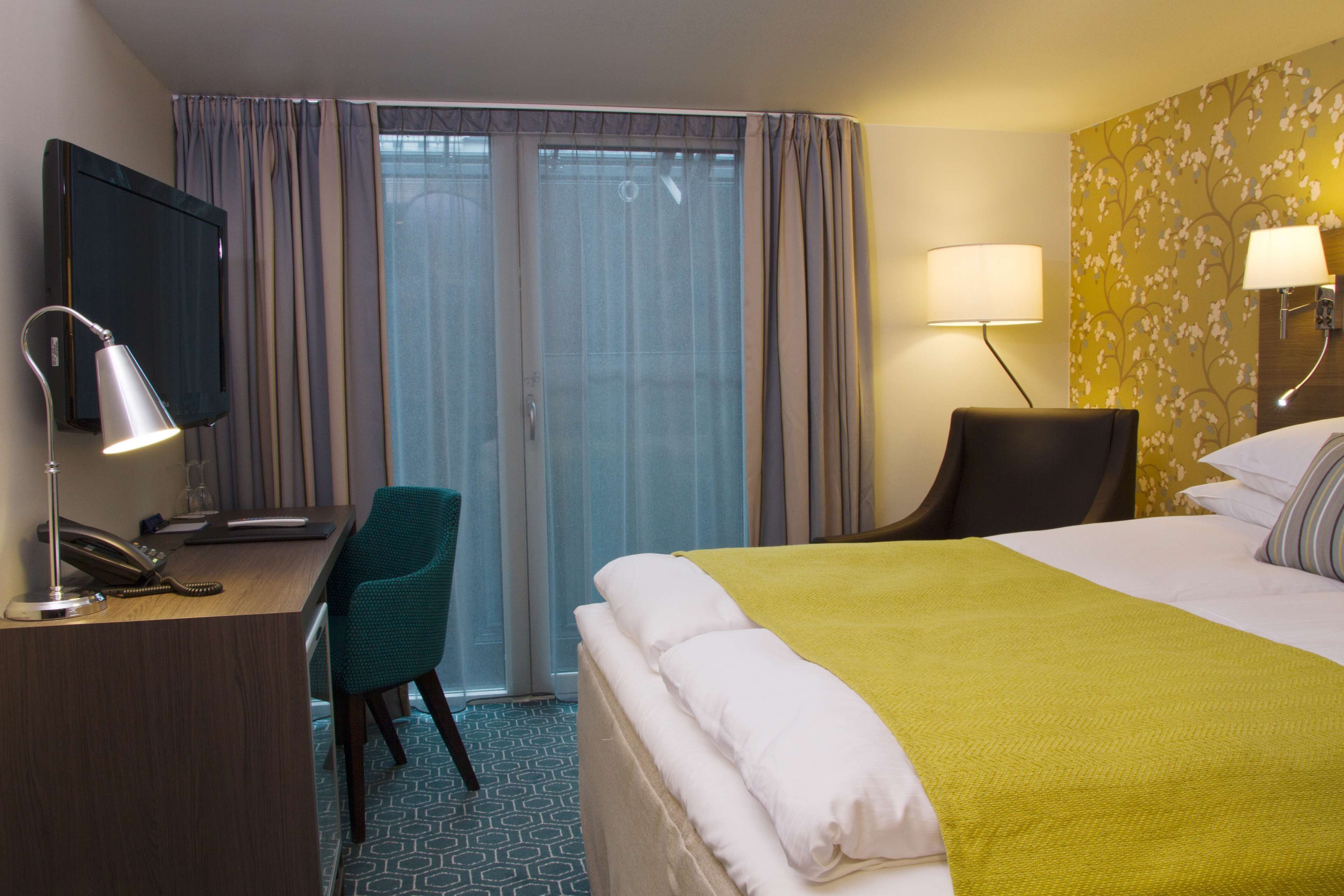 A hotel room with a bed, a desk with a telephone, a chair, a television, and two lamps.