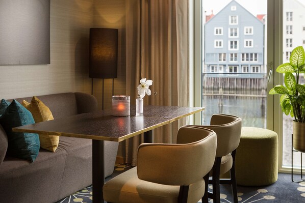 A modern hotel room with a sofa, two armchairs, a small table, a potted plant, and a view of a blue building through large windows.