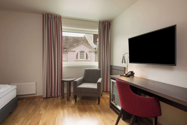A hotel room with a bed, a desk with a computer, a chair, a TV, and a window with curtains.