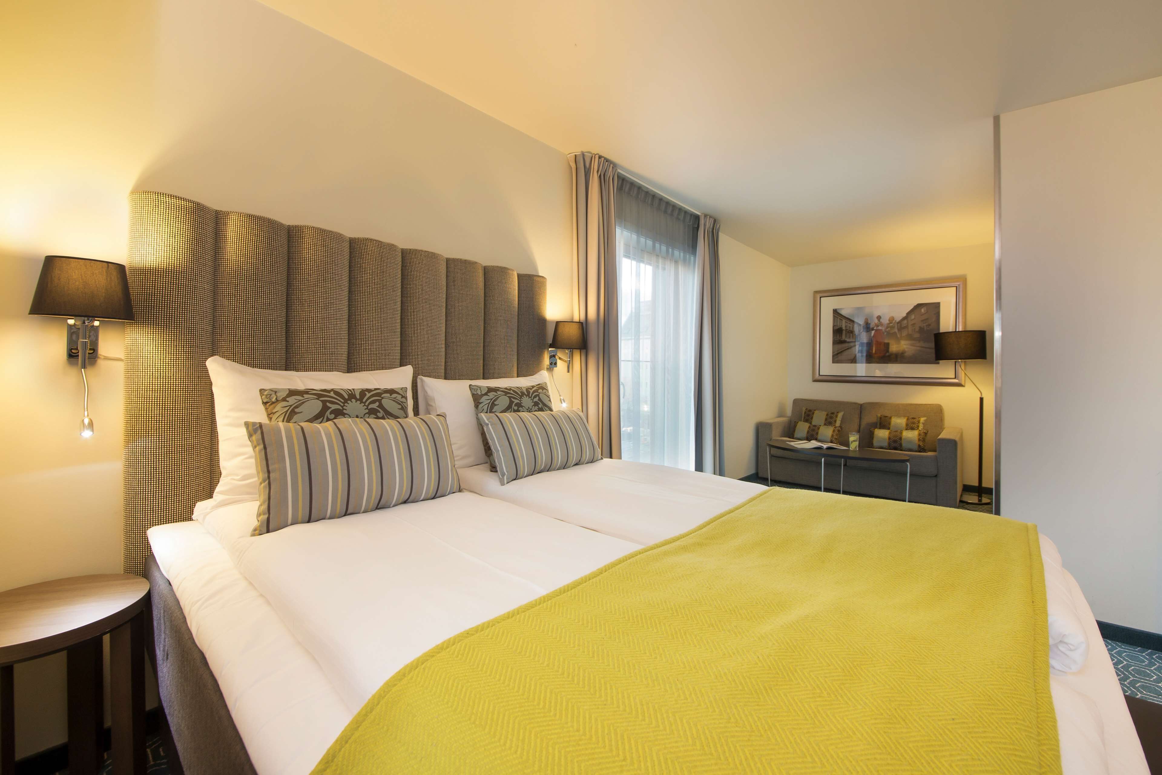 Habitación de hotel con una cama grande, dos mesitas de noche, un sofá y una ventana con cortinas.