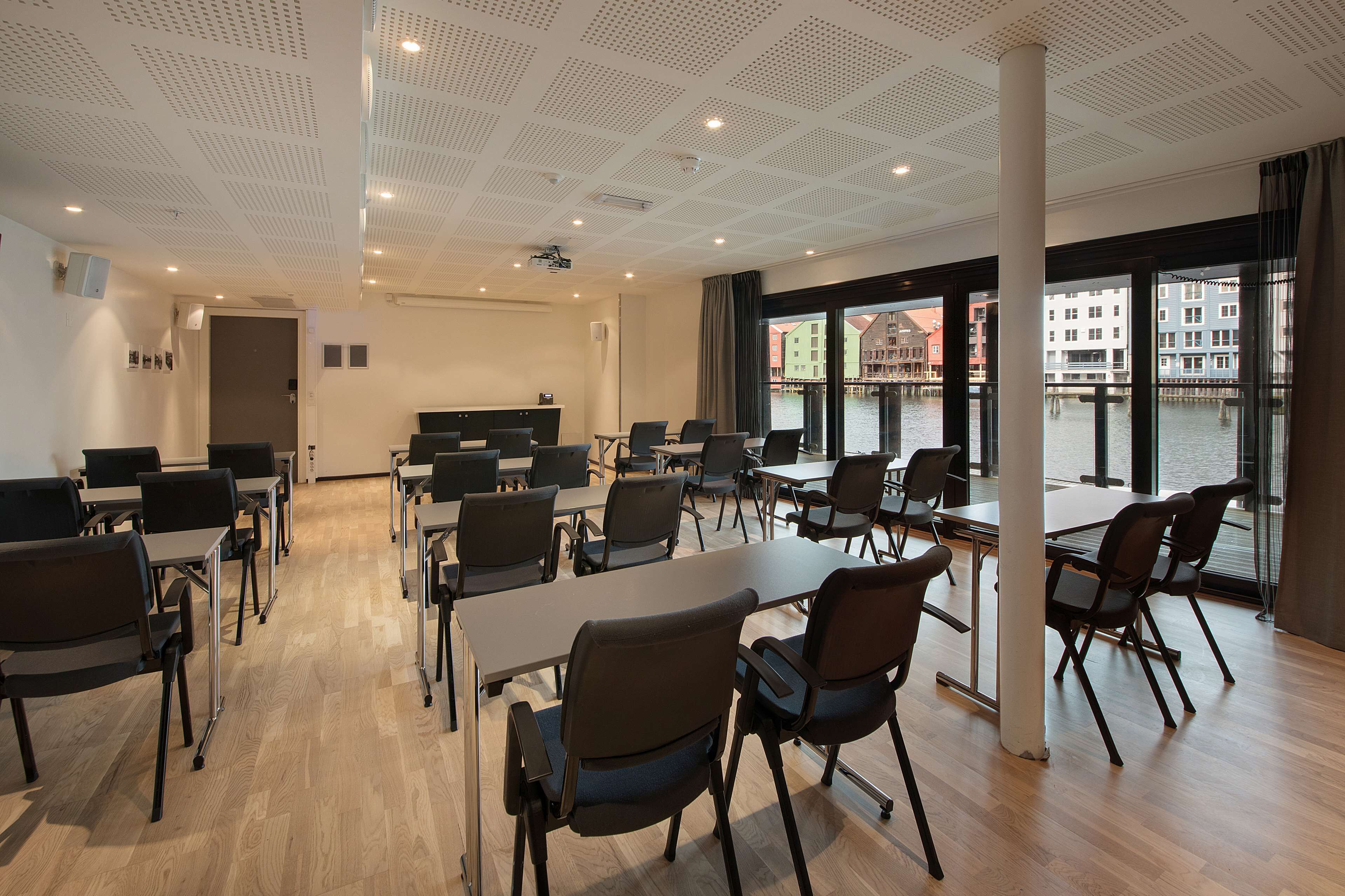 Uma sala de conferências com mesas e cadeiras, grandes janelas com vista para edifícios e para a água, e um teto com padrão de grade.