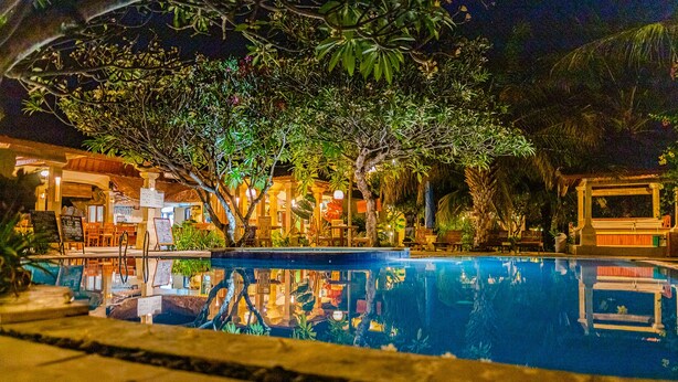 A nighttime view of a hotel with a swimming pool, outdoor seating, and lush greenery.