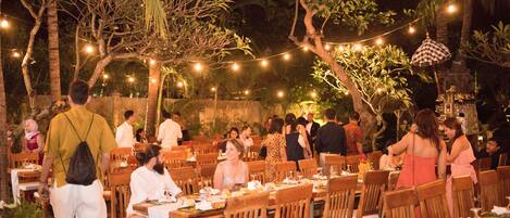 Un espacio exterior para comer con mesas y sillas de madera, rodeado de árboles y luces colgantes.
