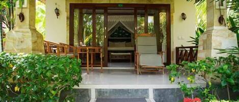 A covered outdoor area with a wooden bench and a small table, surrounded by lush greenery and steps leading to a door.
