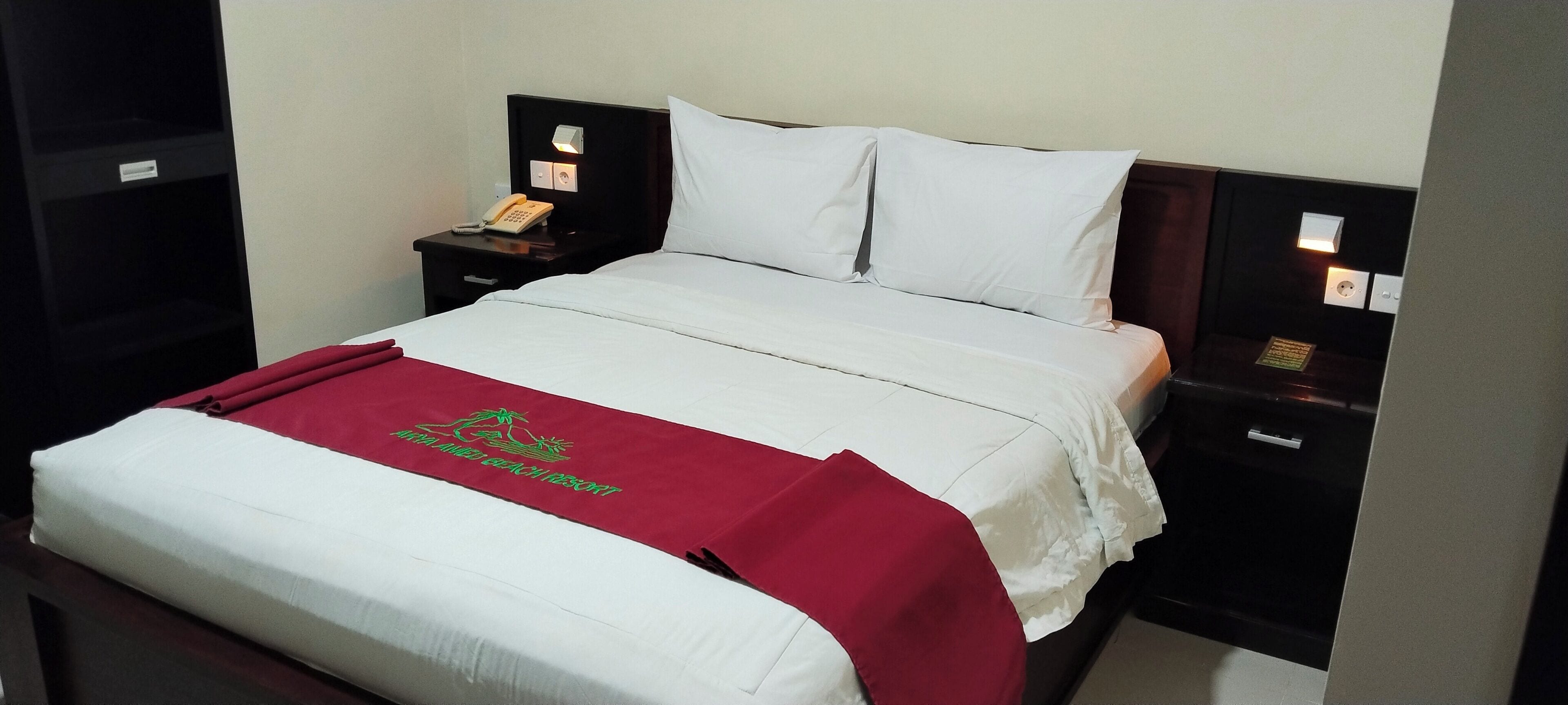 Uma cama bem arrumada com lençóis brancos e um cobertor vermelho, um telefone ao lado da cama e uma pequena mesa de cabeceira com uma luminária.