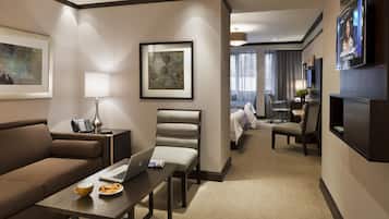 A modern hotel room with a sofa, a desk with a laptop, a dining table with a plate of food, a TV, and a bed in the background.
