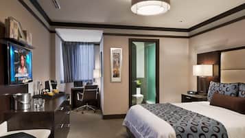 Habitación de hotel con cama, escritorio, silla, televisor y baño visible a través de una puerta abierta.