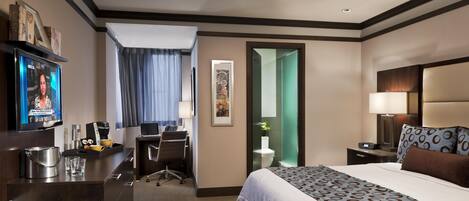 Habitación de hotel con cama, escritorio, silla, televisor y baño visible a través de una puerta abierta.