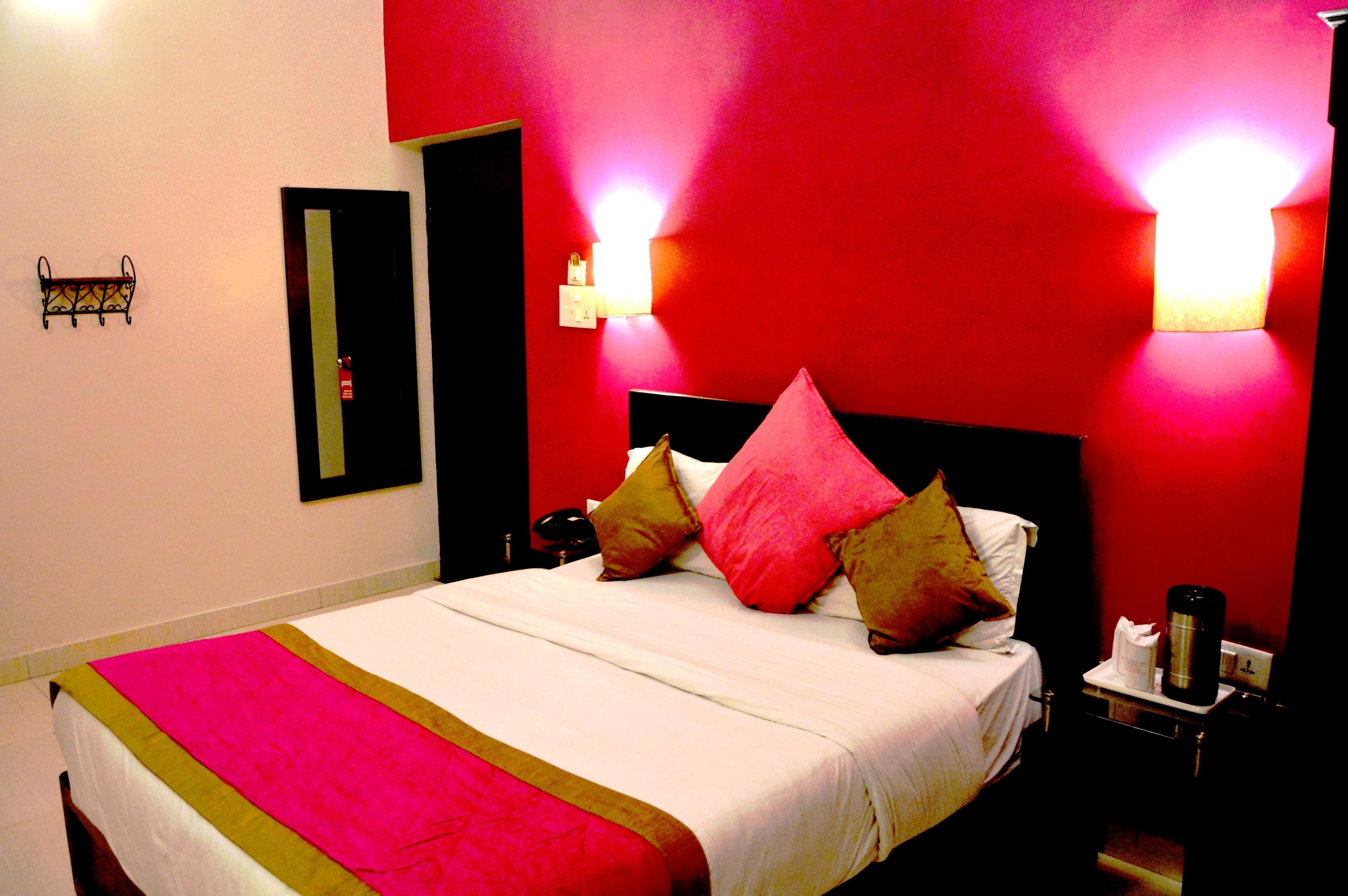 A hotel room with a bed, a nightstand, a red wall, and a mirror.