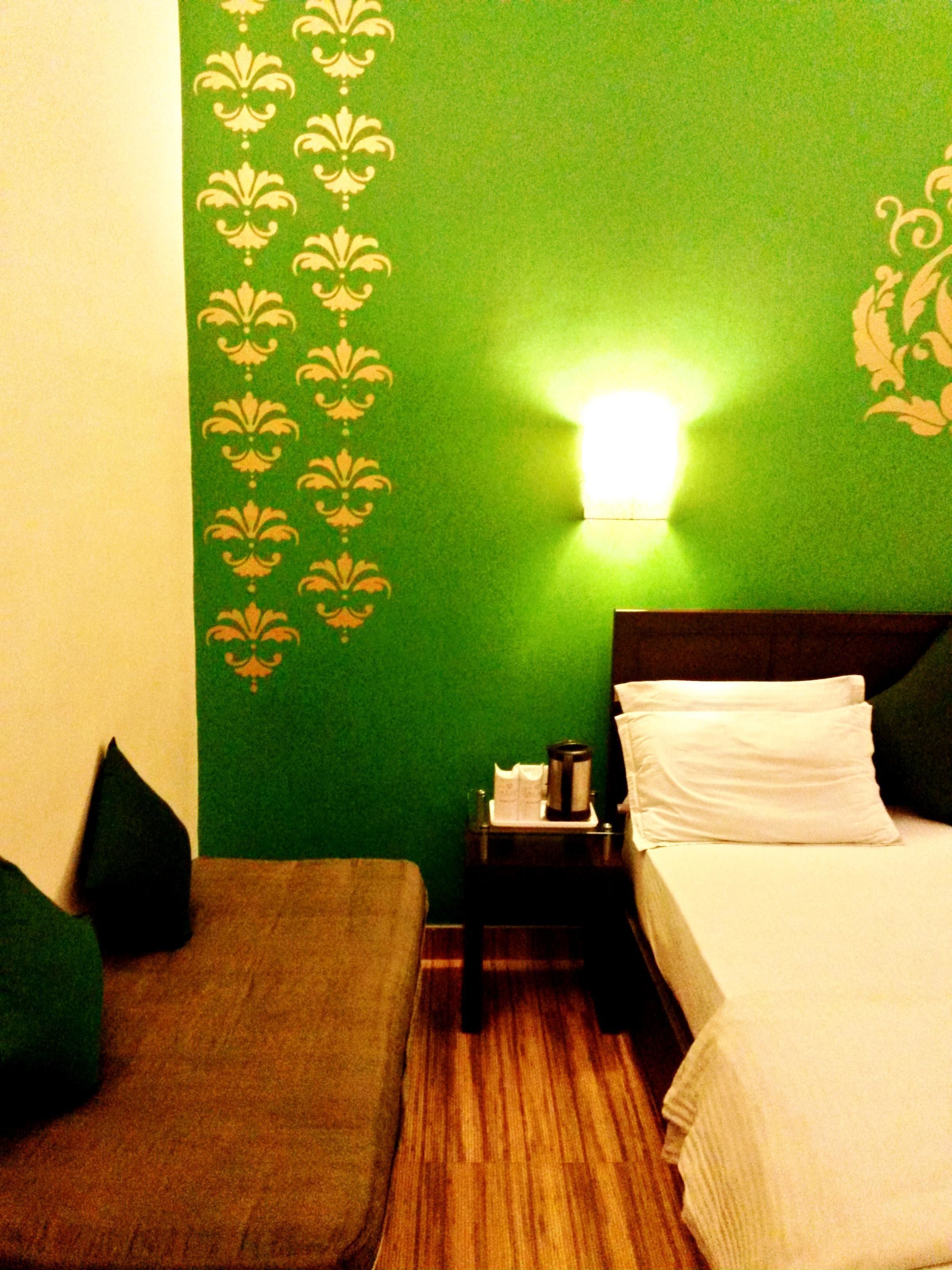 A hotel room with a green wall, a wooden floor, a bed with white bedding, a nightstand with a lamp, and a green cushion on the floor.