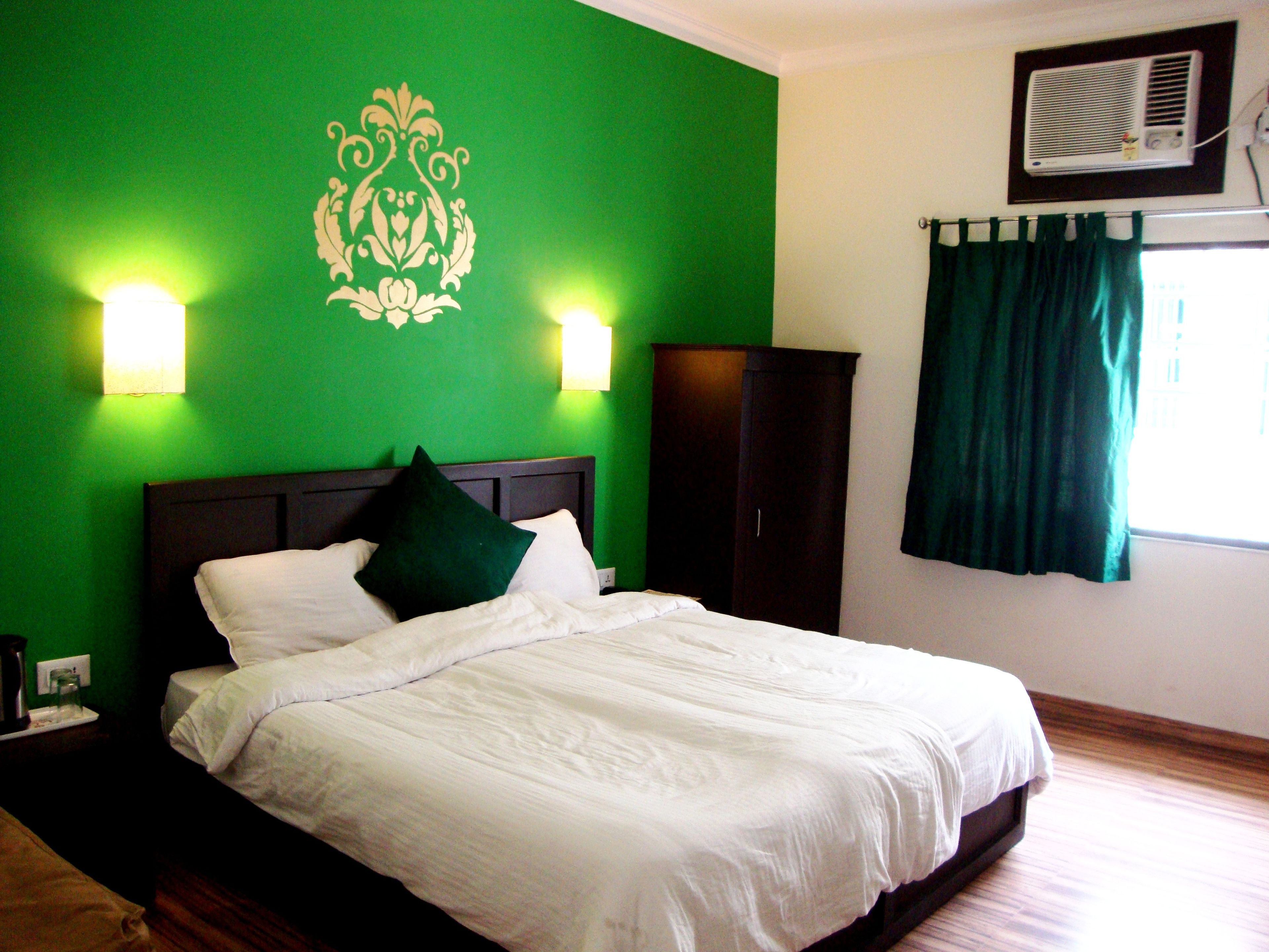 Una cama bien hecha con sábanas blancas y una almohada verde en una habitación con paredes verdes, un armario de madera oscura y una ventana con cortinas verdes.
