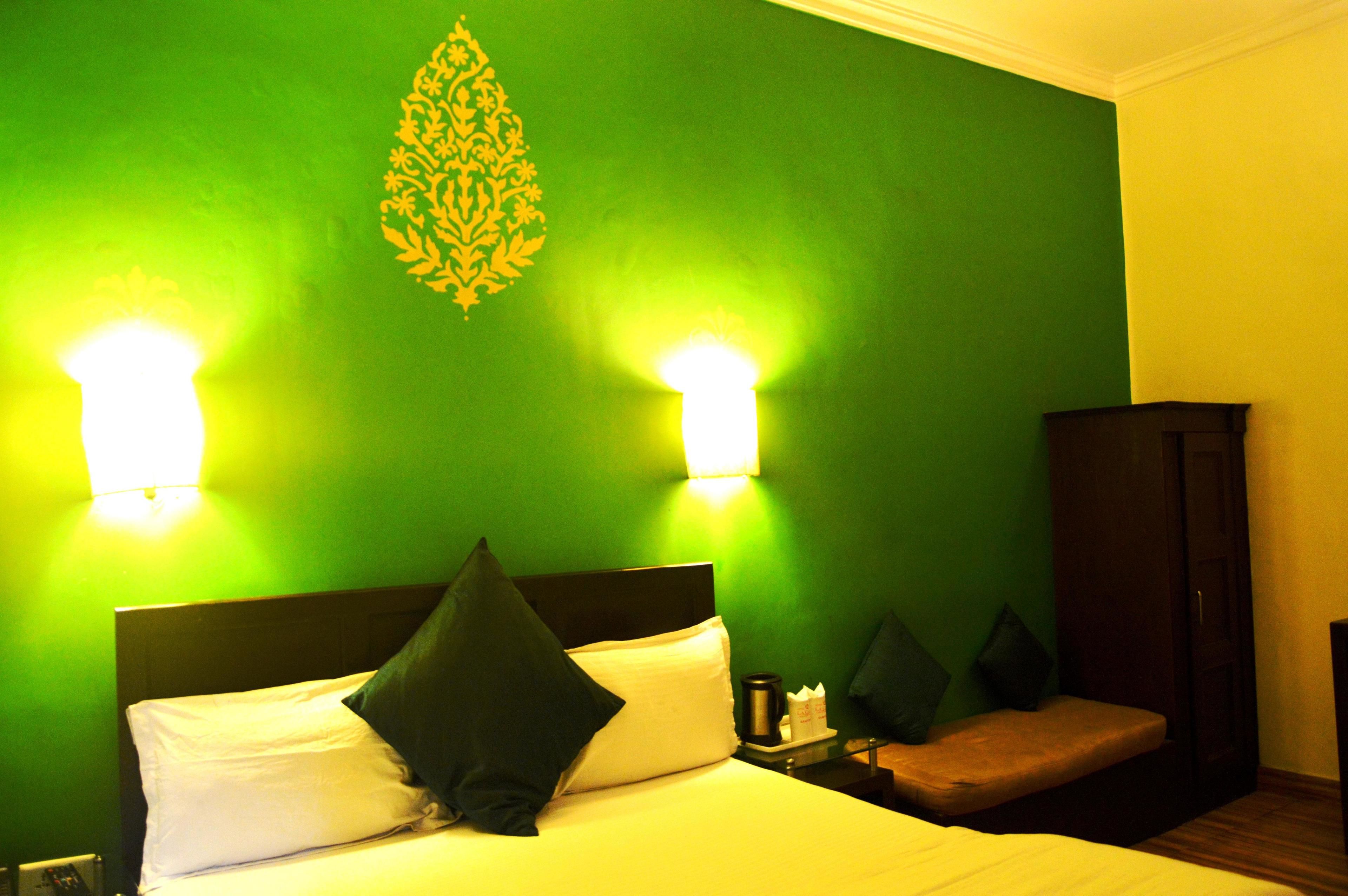 Una habitación de hotel con pared verde, una cama con almohadas blancas y verde oscuro, dos lámparas de noche, una mesita de noche con una cafetera y un armario de madera.