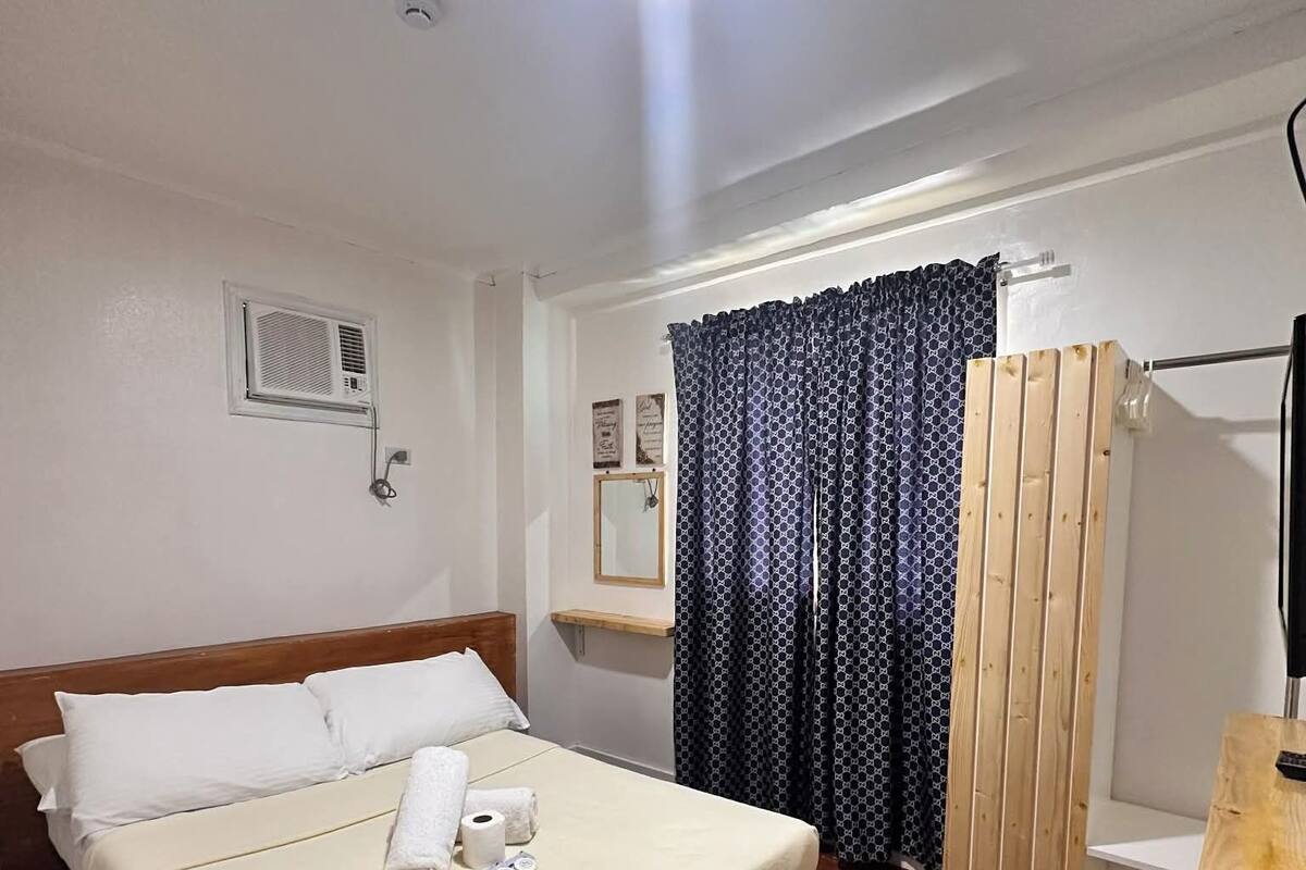 Une chambre d’hôtel avec un lit en bois, une petite table de chevet, un miroir, une fenêtre avec des rideaux à motifs et une cloison en bois.