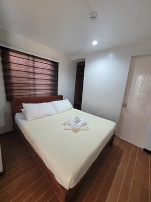 Une chambre d’hôtel avec un lit, une tête de lit en bois, une fenêtre avec des stores, une porte et un luminaire au plafond.