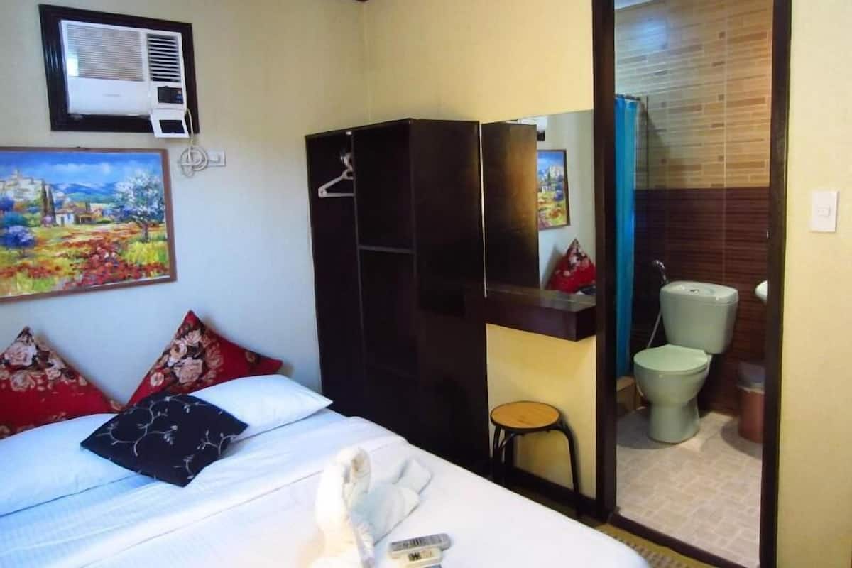 Une chambre d’hôtel comprenant un lit, une armoire, un petit tabouret, un tableau et une salle de bain visible à travers une porte.