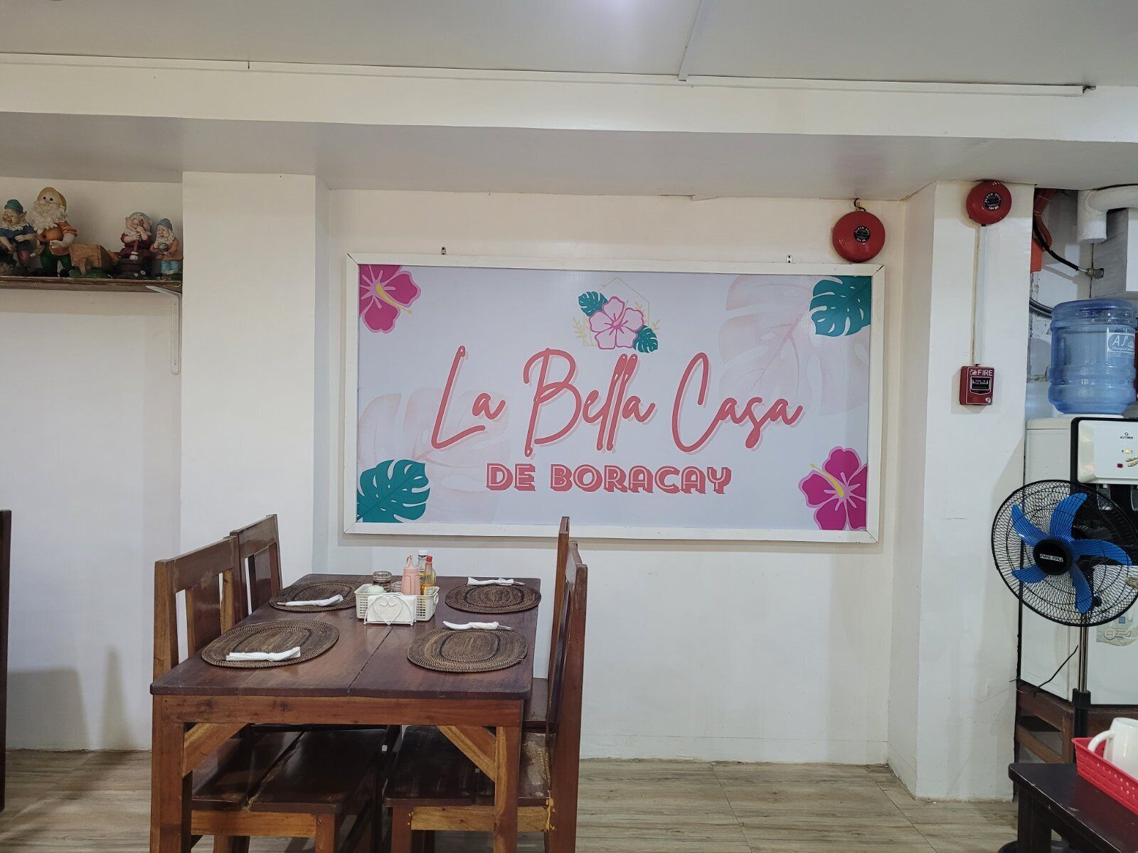 Ein Essbereich mit einem Holztisch, gedeckt für vier Personen, einem an der Wand befestigten Schild mit der Aufschrift 'La Bella Casa de Boracay' und einem Wasserspender auf der rechten Seite.